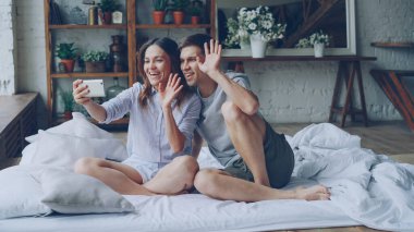 Sevimli Genç çift smartphone ile görüntülü görüşme yapıyor, gençler konuşuyor ve çift kişilik yatak yatak odasında otururken el hareketi. Modern teknoloji ve iletişim kavramı.