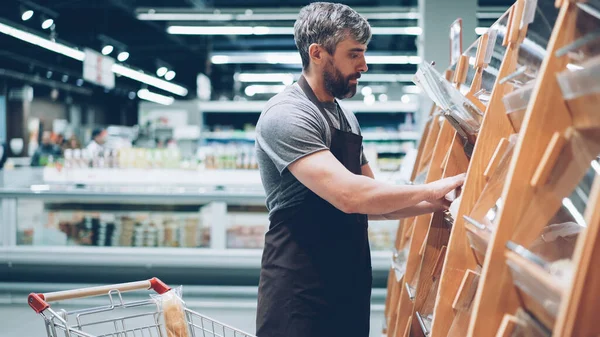 忙碌的男售货员正把面包放在食品店的面包店的货架上 胡子的家伙穿着围裙 销售产品 行业和贸易理念 — 图库照片
