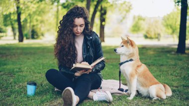 Genç ve güzel kadın parkta kitap okuyor ve sevimli köpek shiba Inu doğurmak okşamaya, iyi yetişmiş evde beslenen hayvan sahibi yakınındaki çimlerde oturuyor. Hayvanlar, Doğa ve insan kavramı.