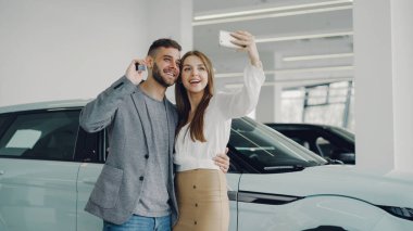 Çekici genç çift selfie arabanın anahtarlarını modern motor showroom ile yeni otomobil satın alma sonra alıyor. Bunlar kameraya bakarak, poz, Öpüşme ve gülümseyen.