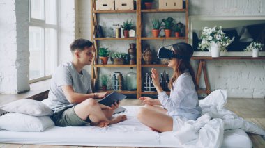 Genç kadın sevgilisi laptop perde dokunmadan, kız arıyorum ve yatağın üstüne oturan gülüyor hareket el ve vücut, sanal gerçeklik gözlük kullanıyor.