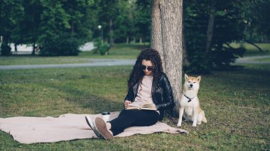 Rahat genç kız kitap okuma ve onun sevimli köpek toprak kokulu iken park ve ağacın altında oturup hava çimenlikte oturan paket servisi olan restoran kahve içme. Eğlence ve Evcil hayvan kavramı.