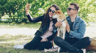 Mutlu çift ile onların köpek çim parkta oturan smartphone kullanarak selfie alıyor, insanlar poz, söz ve shiba Inu köpek yavrusu sevişme. Fotoğraf ve hayvanlar kavramı.