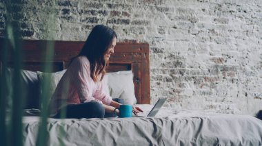 Çekici genç kadın rahat kıyafetler içinde dizüstü bilgisayarda yazarak ve gülümseyen güzel çatı stili yatak odalı evde yatakta otururken. Modern teknoloji ve millennials kavramı.