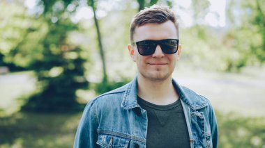 Neşeli genç adam kameraya bakarak ve gülümseyen trendy denim ceket giyen güneş gözlüğü yakın çekim portresi. Güneşli yaz gün, arka planda güzel yeşil park.