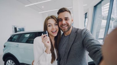 Çekici genç çift selfie arabanın anahtarlarını modern motor showroom ile yeni otomobil satın alma sonra alıyor. Bunlar kameraya bakarak, poz, Öpüşme ve gülümseyen.