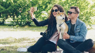 Mutlu çift ile onların köpek çim parkta oturan smartphone kullanarak selfie alıyor, insanlar poz, söz ve shiba Inu köpek yavrusu sevişme. Fotoğraf ve hayvanlar kavramı.