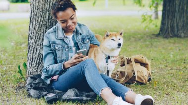 Mutlu Afrikalı-Amerikalı kadın modern smartphone kullanarak ve onun sevimli hayvan köpek şehir parkında rüzgarlı yaz günü dinlenme okşama. Doğa, hayvanlar ve insanlar kavramı.