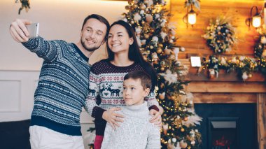 Sevgi dolu baba, anne ve oğluyla yeni yıl ağacının yanında duran akıllı telefon kamerasıyla selfie çekiyor. Fotoğraflar, insanlar ve aile tatilleri konsepti.