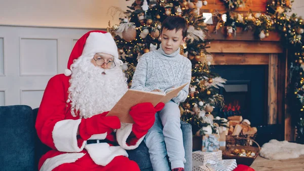 サンタクロースはクリスマスの日に装飾された家のソファに座って子供を祝う幸せな男の子に本を読んでいます 幼少期と文学の概念 — ストック写真