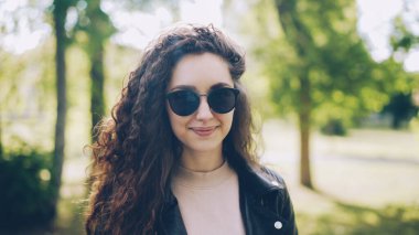 Portre çekici genç kadın güneş gözlüğü ve trendy deri ceket kameraya bakarak ve şehir parkı önünde duran yaz gününde gülümseyen. İnsanlar ve güneş ışığı kavramı.