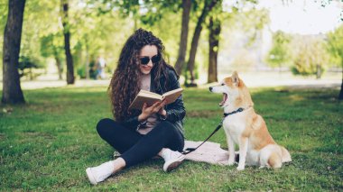 Neşeli kız öğrenci parkta kitap okuyor çimenlerde oturuyor ve güzel köpeği okşuyor, evcil hayvan esniyor kız gülüyor..