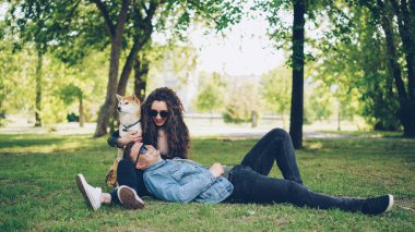 Yakışıklı genç kız arkadaşı onunla konuşuyor ve sevimli köpek sevişme, Çift gülüyor ve yaz hafta sonu tadını parkta çim üzerinde yatıyor.