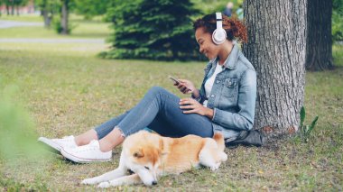 Çekici Afrikalı-Amerikalı kadın ve onu safkan köpek çim yeme yakın yatarken çim parkta oturan smartphone kullanarak bir kulaklık ile müzik dinlemek.