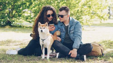 Neşeli karı koca Park, çim üzerinde dinlenme smartphone ekran ve konuşan ve Füssing köpek izlerken kullanıyorsunuz. Modern teknoloji, Evcil hayvanlar ve şehir kavramı.