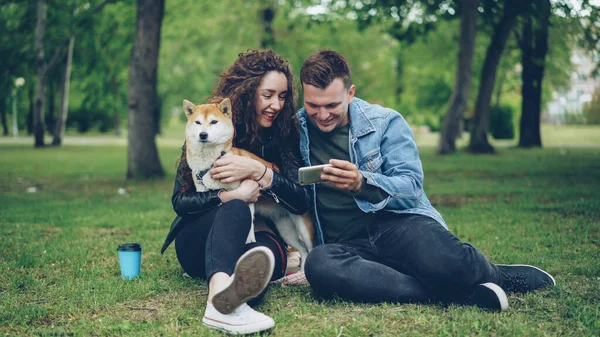 Sevgi dolu erkek genç ve güzel kadın tutarak ve şehir Park'ın bahçesinde otururken iyi yetişmiş köpeği okşamaya için smartphone ekran gösteriyor. İnsanlar ve modern teknoloji kavramı.
