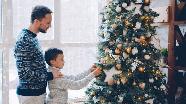 Baba ve oğul yeni yıl ağacını süslüyorlar parlak toplar asıyorlar ve evlerinde modern rahat evlerinde konuşuyorlar. Aile, insanlar ve kutlamalar konsepti.