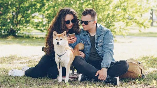 Neşeli karı koca Park, çim üzerinde dinlenme smartphone ekran ve konuşan ve Füssing köpek izlerken kullanıyorsunuz. Modern teknoloji, Evcil hayvanlar ve şehir kavramı.