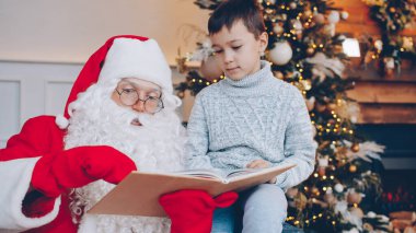 Sevimli çocuk ve Noel Baba Noel ağacının yanında kitap okuyor ve yılbaşı günü konuşuyorlar. Çocukluk, tatil ve mutluluk kavramı.