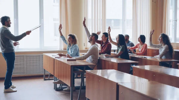 フレンドリーな先生は 若い人が手を上げている間質問をし答えるデスクに座って陽気な学生に話しています 若者の概念 — ストック写真