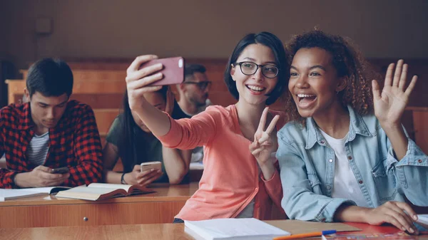 学生は大学でテーブルに座ってのスマート フォン Selfie を取っている元気な女の子 若い女性がポーズをとって手ジェスチャーを作ると抱き締めます 友人や技術コンセプト — ストック写真