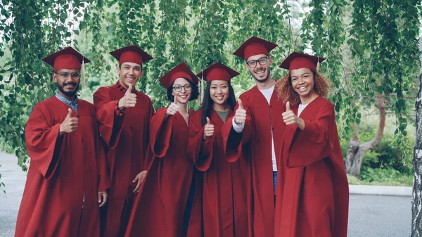 Portrait of excited graduating students multiethnic group standing outdoors in red gowns showing thumbs-up and looking at camera.