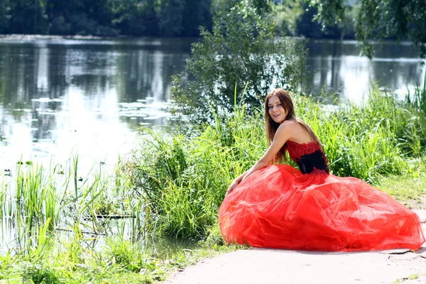 Nehrin kenarında oturan kırmızı elbiseli, gülümseyen kadın. — Stok fotoğraf