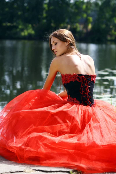 Mujer sonriente en un vestido rojo sentado cerca del río — Foto de Stock