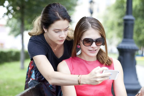 Smiling young girls with cell phone sitting on a bench in a park — Stock Photo, Image