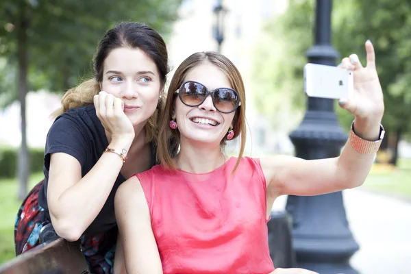 Chicas jóvenes sonrientes con teléfono celular sentadas en un banco en un parque — Foto de Stock