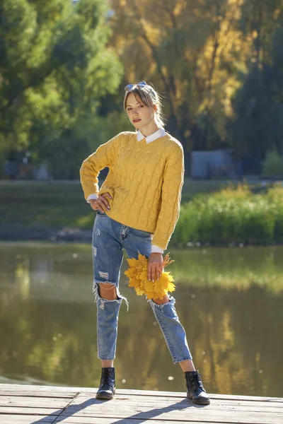 秋の公園でポーズをとるウールのセーターと引き裂かれたジーンズの若い女の子の完全な長さの肖像画 — ストック写真
