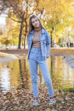 Güz parkında poz veren şık, yırtık pantolonlu güzel bir kızın tam boy portresi.