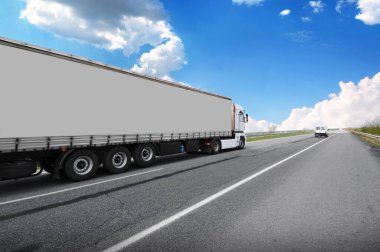 Big truck with a grey trailer with a space for text on a countryside road with other cars against a blue sky with clouds