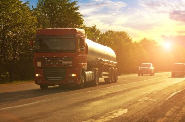 Büyük kırmızı tanker diğer arabalarla kırsal bir yolda yeşil ağaçlarla birlikte gün batımına karşı
