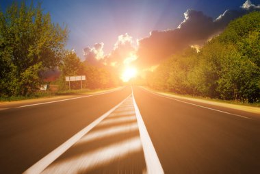 Yaz kırsalında asfalt yol, yeşil ağaçlarla birlikte, parlak günbatımına sahip mavi gökyüzüne karşı hareket ediyor.