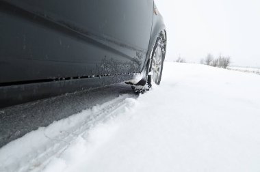 Close-up of a black car on a winter countryside road covered in snow against a white sky