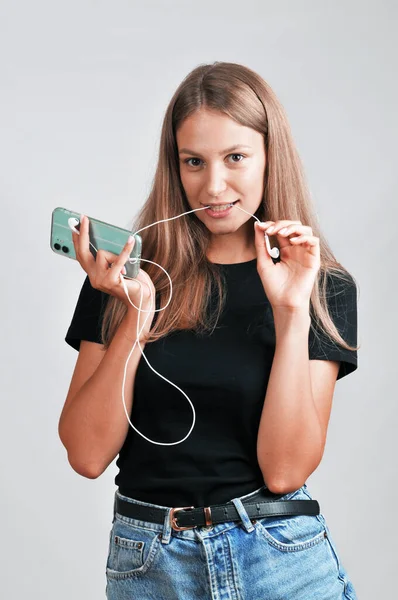 一个穿着黑色T恤和牛仔裤短裤 拿着智能手机 带着灰色背景的咬着耳机的年轻女子的画像 — 图库照片