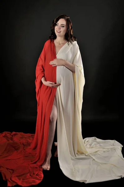 穿着浅色米黄色衣服和红色衣服的漂亮孕妇在黑色背景上显得孤立无援 免版税图库照片
