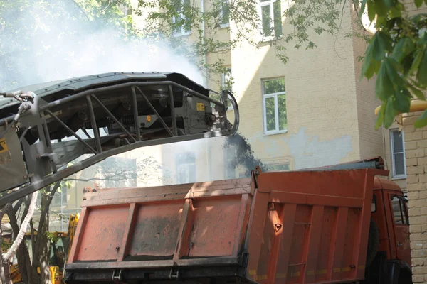 Road milling vehicle removes old asphalt and loads milled asphalt into dump truck inside a narrow courtyard with old houses. Road repair works.