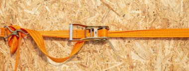 Ratchet truck cargo tie downs clasp chipboard box. Orange load belt over wooden osb surface. clipart
