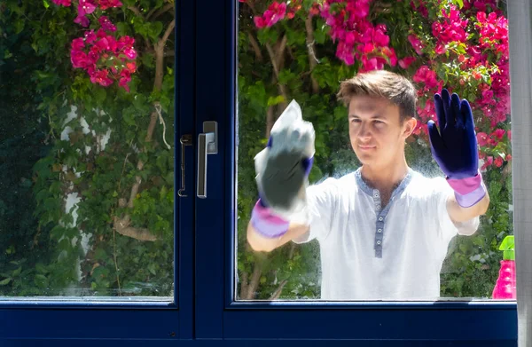 Handsome guy in gloves cleaning window with rag and detergent at home. House cleaning. Washing dirty window glass. Housework and housekeeping concept