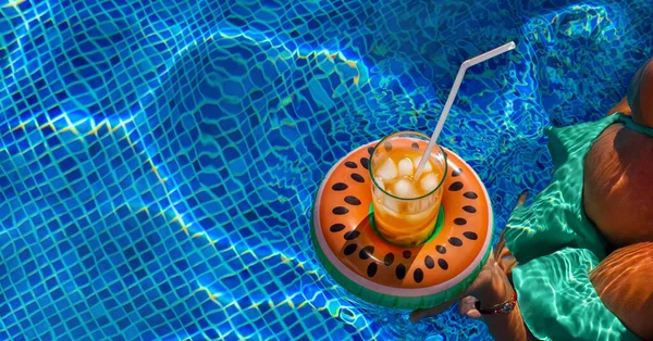 Cold refreshing drink with ice and a straw in the inflatable ring in shape of watermelon in a woman\'s hand in a pool of blue water. Concept of summer holiday in hotel or pool party. Copy space, banner