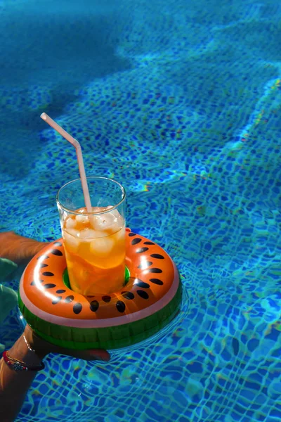 Cold refreshing drink with ice and a straw in the inflatable ring in shape of watermelon in a woman\'s hand in a pool of blue water. Concept of summer holiday in hotel or pool party. Copy space