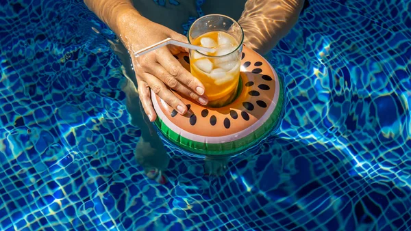 Cold refreshing drink with ice and a straw in the inflatable ring in shape of watermelon in a woman\'s hand in a pool of blue water. Concept of summer holiday in hotel or pool party. Copy space