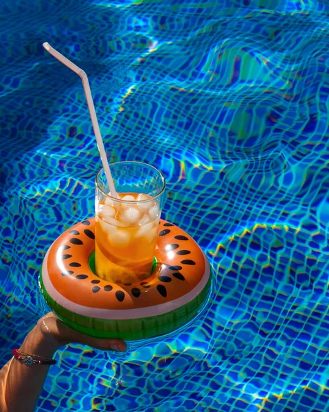 Cold refreshing drink with ice and a straw in the inflatable ring in shape of watermelon in a woman\'s hand in a pool of blue water. Concept of summer holiday in hotel or pool party. Copy space