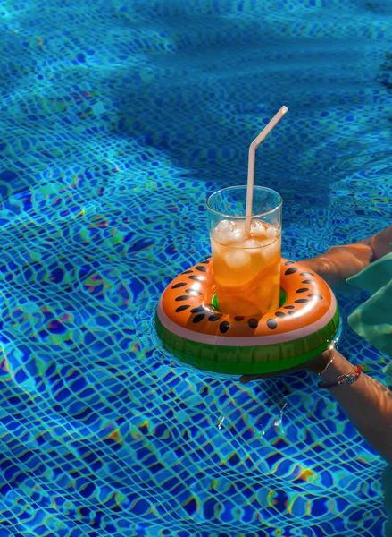 Cold refreshing drink with ice and a straw in the inflatable ring in shape of watermelon in a woman\'s hand in a pool of blue water. Drinks holders for swimming pool. Concept of summer holiday in hotel or pool party. Copy space