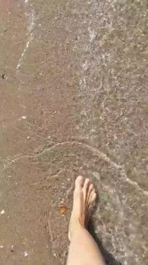 Woman feet walking along sea water waves on sandy beach. Female on summer travel vacation, slow motion. Sitges beach on a sunny day, Barcelona, Spain. Concept of summer vacation