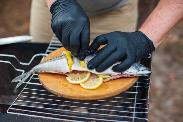 Grilled mackerel fish with lemon slices, top view. The process of cooking fish outdoors. Chef in black latex gloves preparing mackerel fish on a wooden cutting board. Tasty and fresh food, picnic, party, outdoor recreation