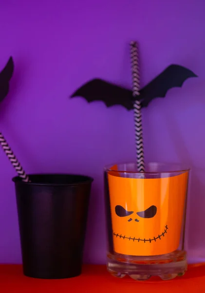 Halloween decor. Paper cup with straw inside and paper silhouettes of bats, bright background. Spooky holiday symbols, selective focus. Space for text