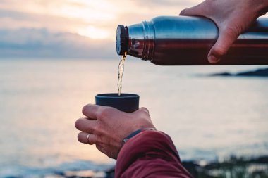A close-up shot of man pouring hot tea from thermos on the background of the ocean on the sunset